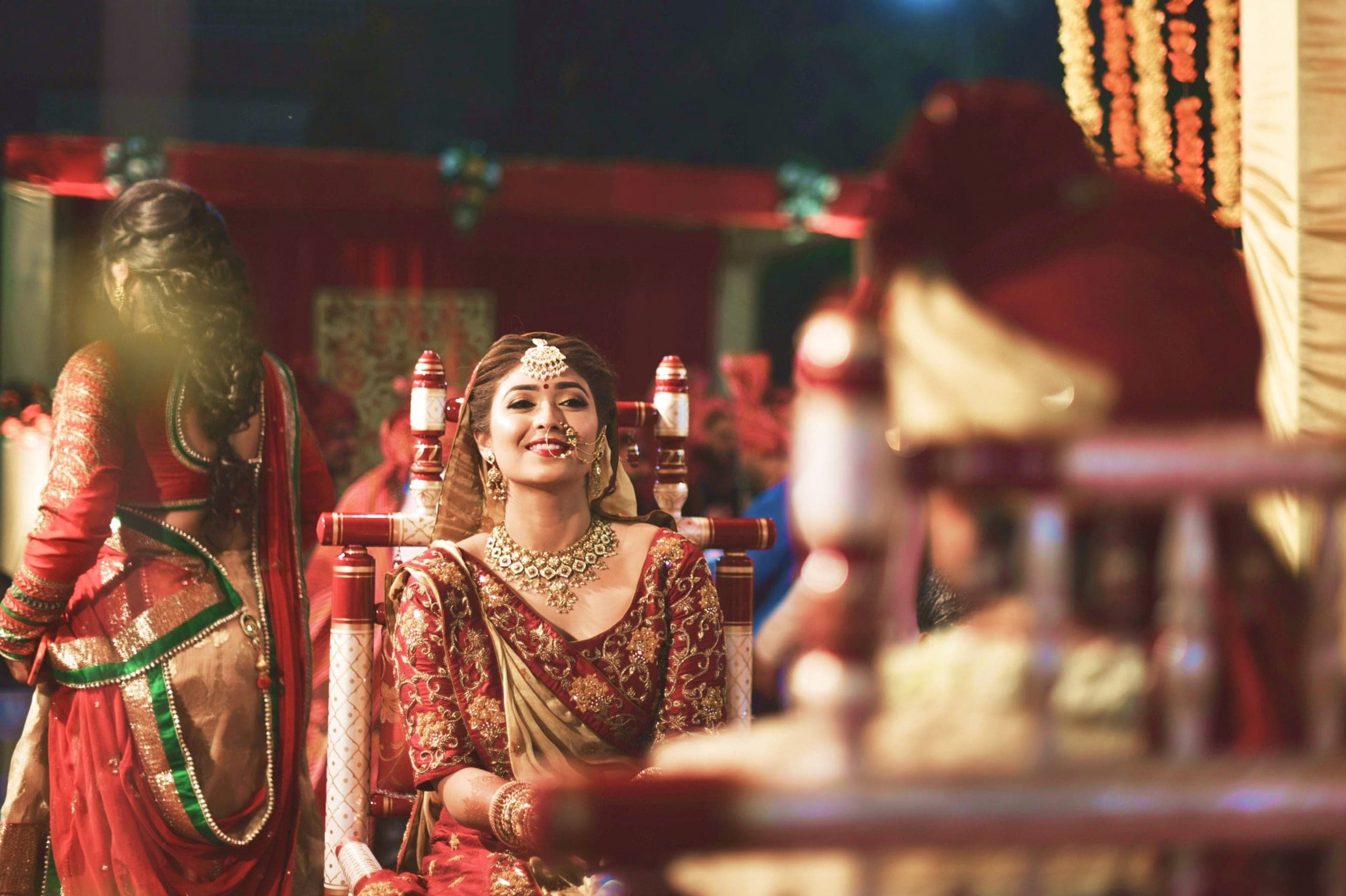 A bride in a red and gold lehenga seated with her hands in her lap during a wedding ceremony.