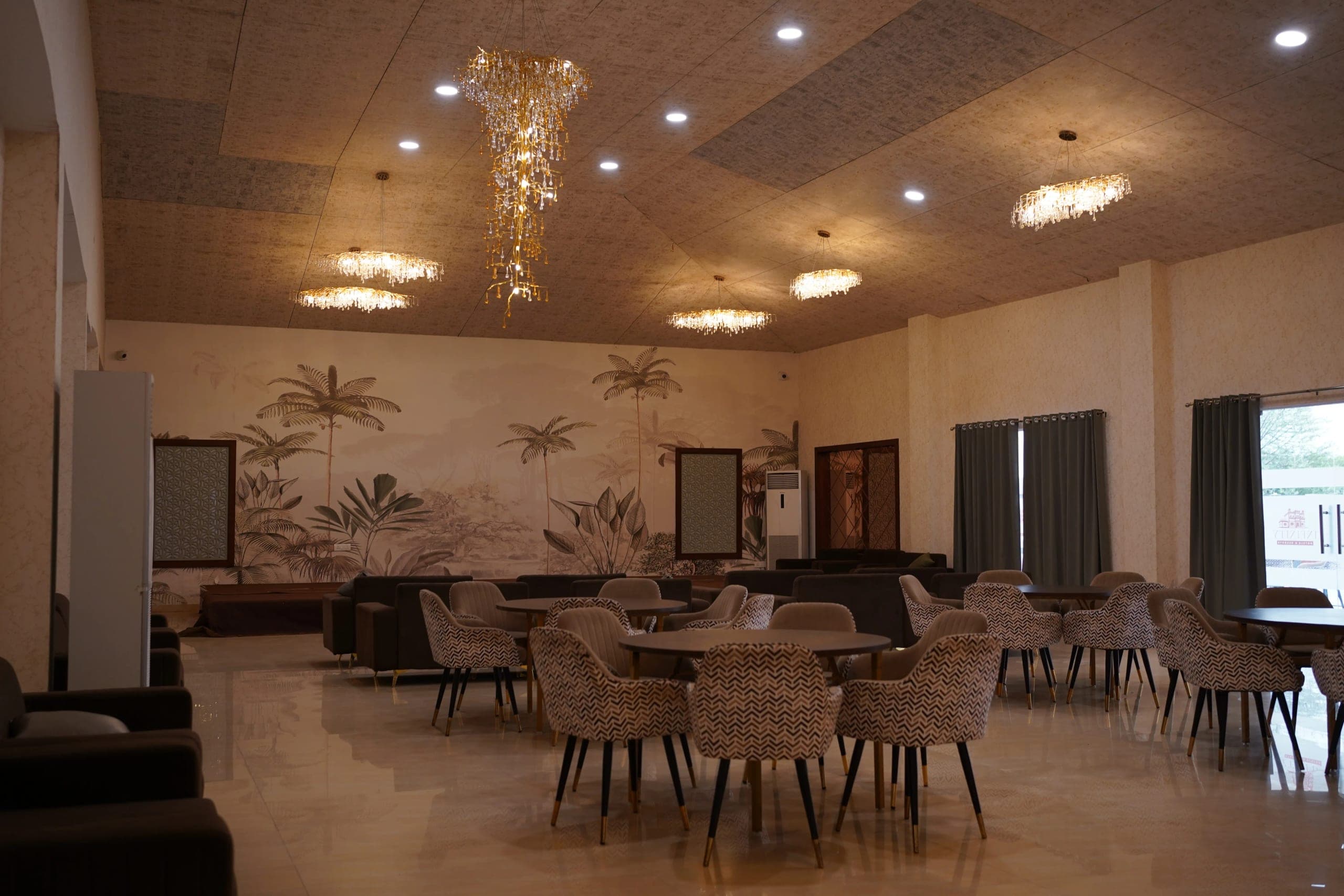 The indoor restaurant: round tables with patterned upholstered armchairs on a polished marble floor, deep sofas along the near wall, tiered crystal chandeliers overhead, and a painted mural of palms and tropical foliage running the length of the far wall.