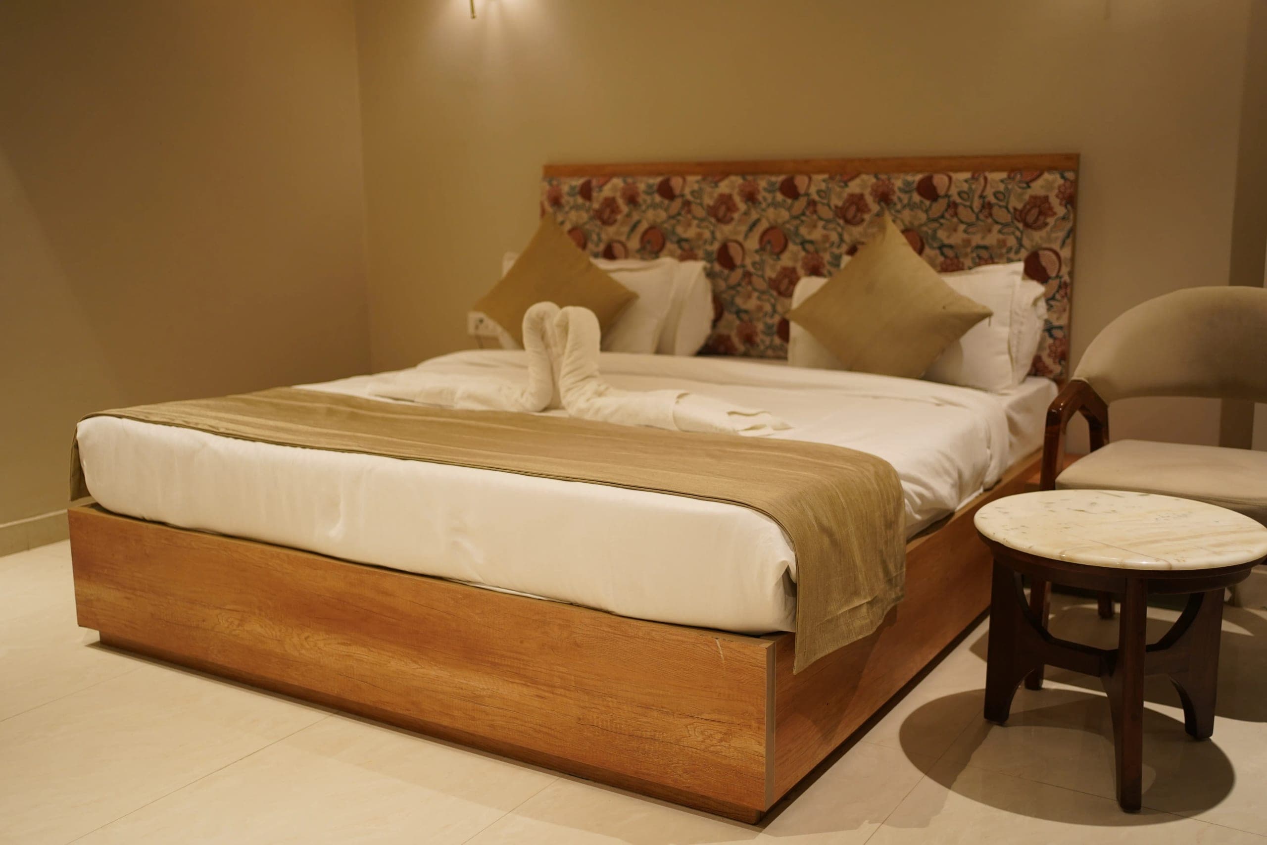 A guest room bed on a pale teak platform base, set against a full-width headboard panel upholstered in block-printed floral fabric, dressed in white linen with a taupe runner and cushions, beside a round marble-topped side table and an armchair.