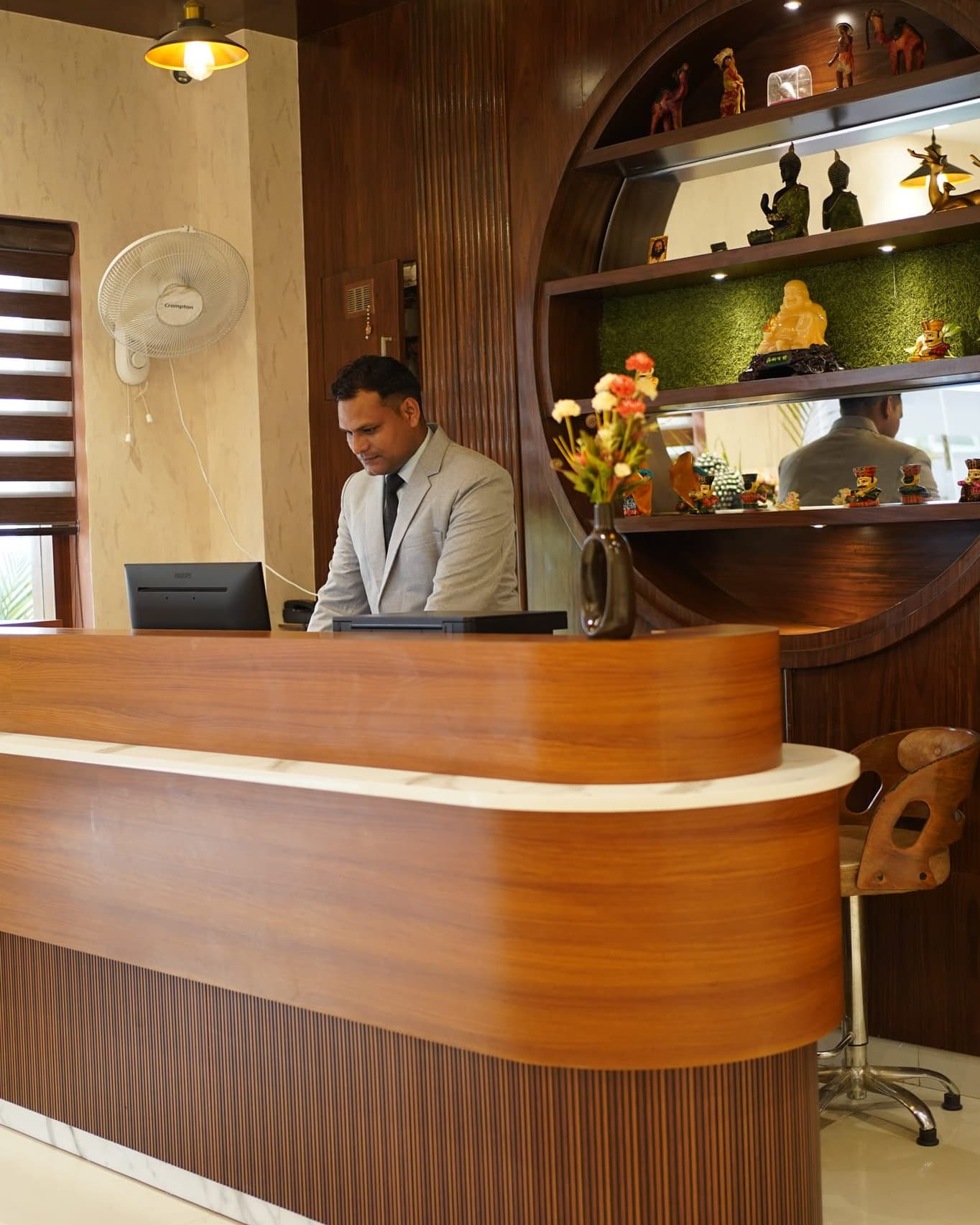 A member of the front-desk team working at the curved teak reception counter, beside a tall circular niche of backlit shelves holding carved wooden curios.
